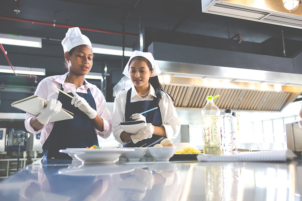 Photo of two kitchen staff making notes on recipes they are observing