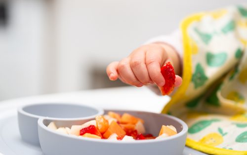 Macro Close Up Of Baby Hand With A Piece Of Fruits Sitting In Child's Chair Kid Eating Healthy Food